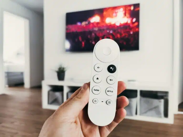 au premier plan, une main qui tient une télécommande blanche avec 8 boutons dont 1 noir, tous ronds. À l'arrière-plan, une tv accrochée au mur, allumée, dans un salon à la décoration moderne.